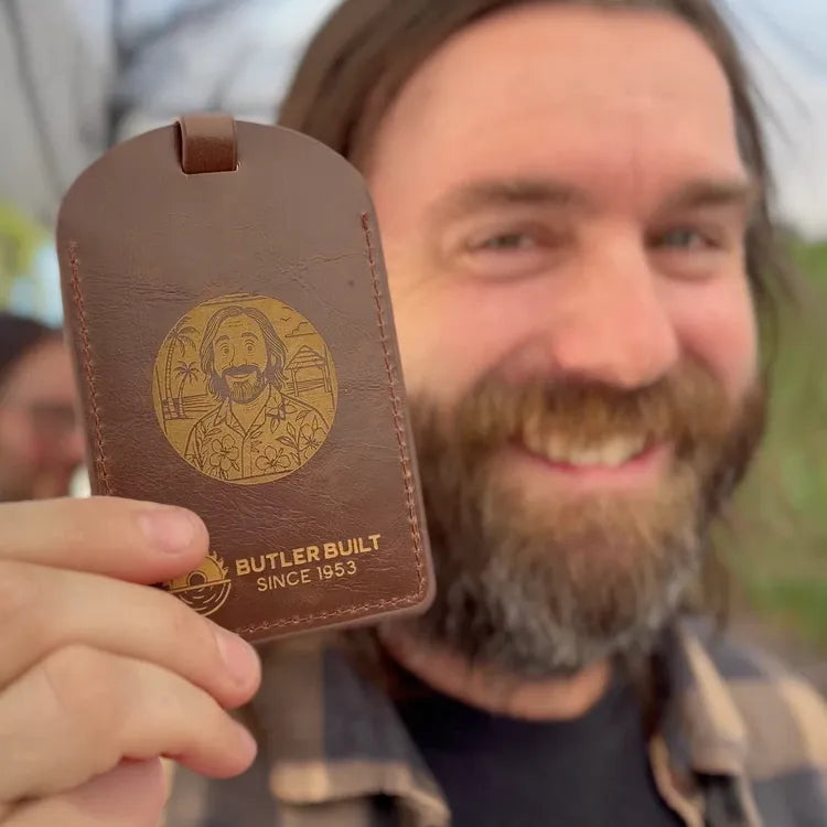 A smiling bearded man holds up a brown leather luggage tag or key fob-style card with a gold-toned engraved portrait and the text 'BUTLER BUILT SINCE 1953' against an outdoor blurred background.