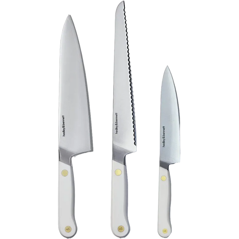 Three Hedley & Bennett stainless steel kitchen knives with white handles and brass rivets, including a chef's knife, a serrated bread knife, and a utility knife.