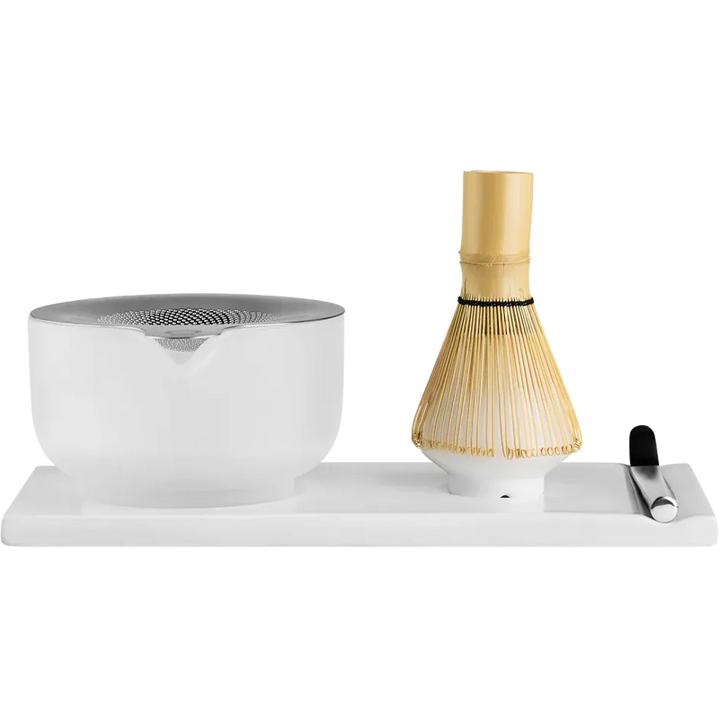 Fellow matcha tea preparation set on a white rectangular base, featuring a frosted white ceramic bowl with a metallic silver lid, a bamboo whisk with a gold-colored handle, and a small black-handled tool on the right.