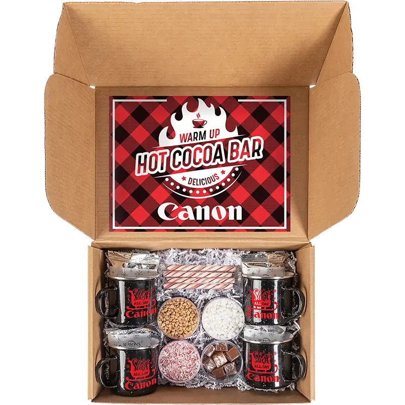 An open cardboard gift box containing a hot cocoa bar set. The lid features a red and black plaid graphic with the text 'WARM UP HOT COCOA BAR' and the brand name 'Canon'.