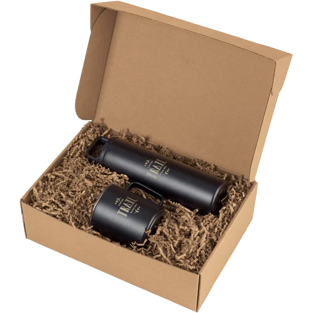 An open kraft cardboard gift box filled with brown crinkle paper, containing two matte black insulated tumblers with gold foil branding that reads 'The Trail'.