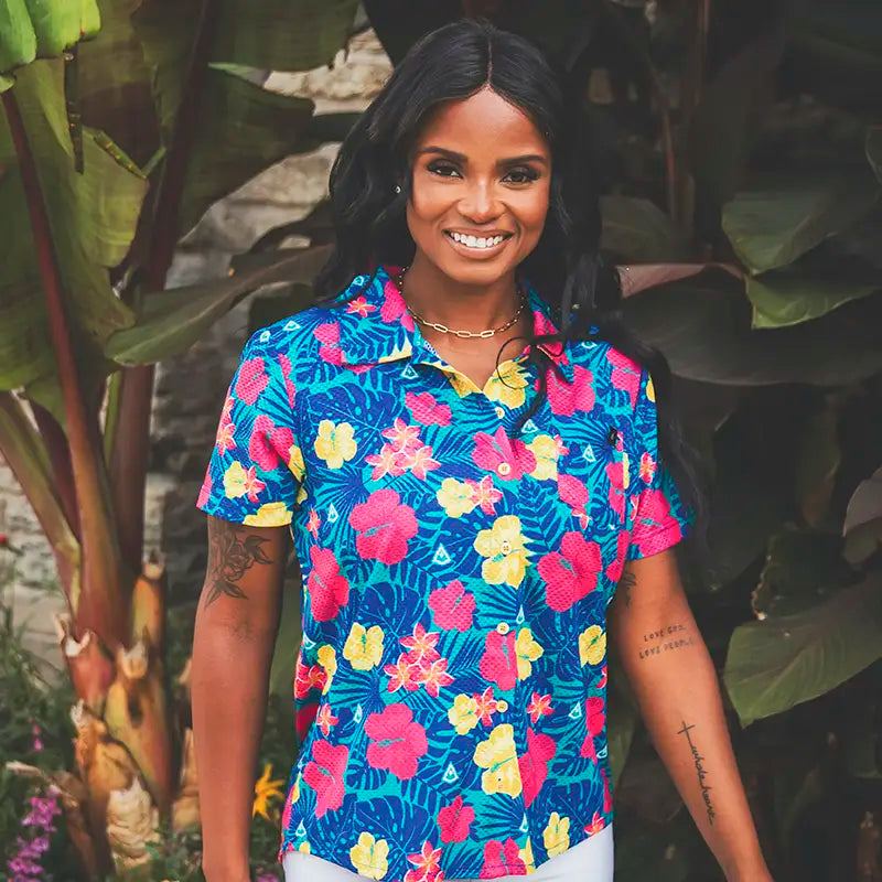 A woman smiling outdoors in front of large tropical green leaves, wearing a short-sleeve collared button-up shirt with a bright tropical floral print in blue, pink, yellow, and green.