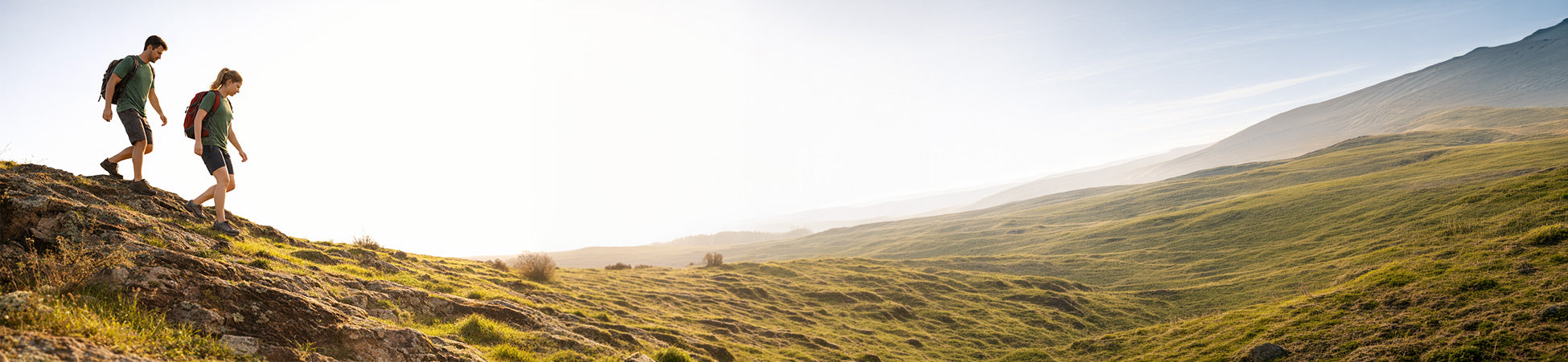 Two hikers walking across a grassy hillside overlooking a mountain valley