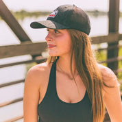 A woman wearing a black trucker-style cap with a subtle tonal wave pattern and a small logo patch on the front, photographed outdoors near water and a wooden railing.