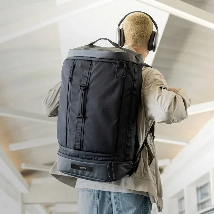 Person wearing a large black backpack and headphones, standing in an indoor setting.