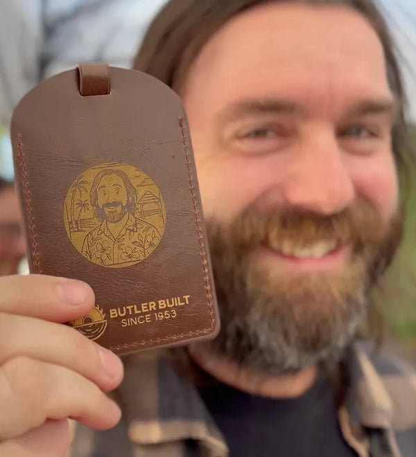A smiling bearded man holds up a brown leather luggage tag or key fob-style card with a gold-toned engraved portrait and the text 'BUTLER BUILT SINCE 1953' against an outdoor blurred background.
