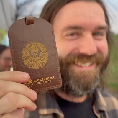 A smiling bearded man holds up a brown leather luggage tag or key fob-style card with a gold-toned engraved portrait and the text 'BUTLER BUILT SINCE 1953' against an outdoor blurred background.