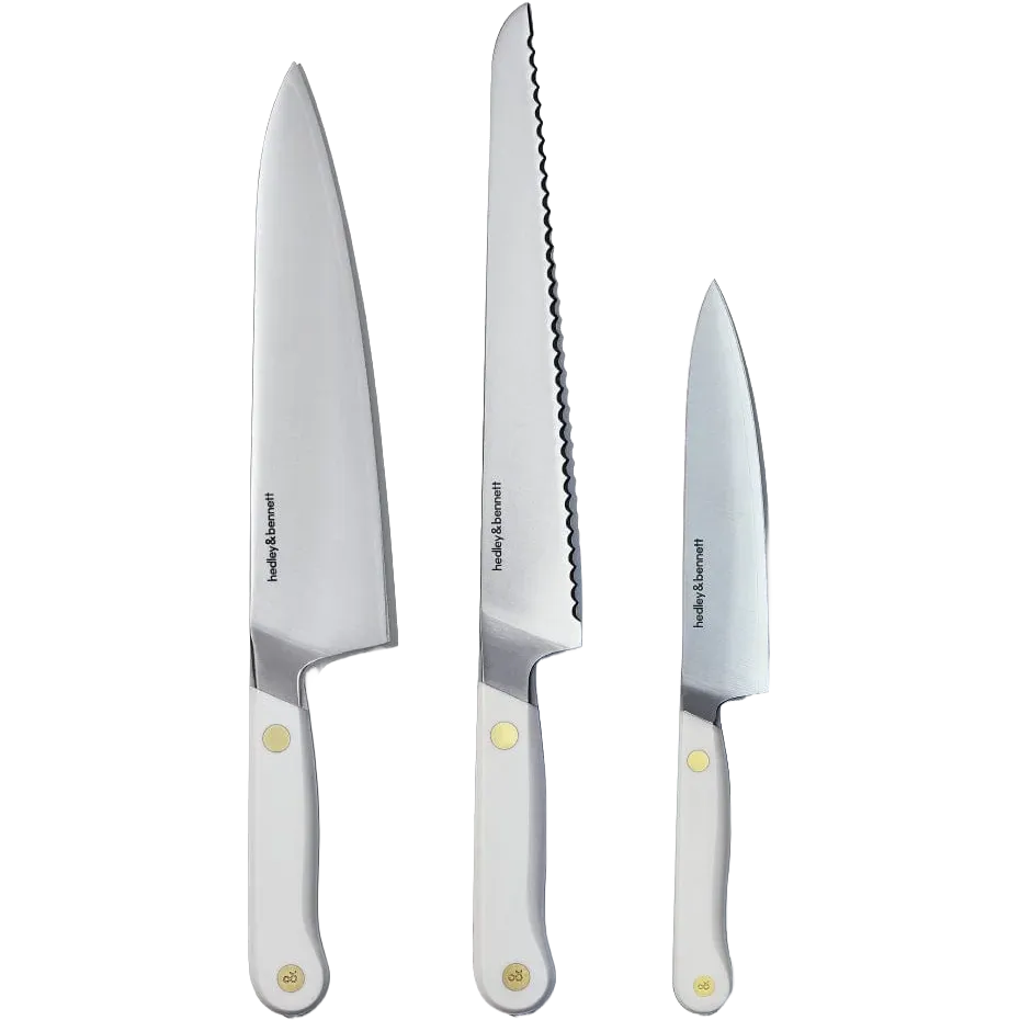 Three Hedley & Bennett stainless steel kitchen knives with white handles and brass rivets, including a chef's knife, a serrated bread knife, and a utility knife.