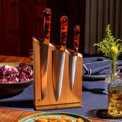 A Hedley & Bennett wooden knife block holding three kitchen knives with tortoiseshell handles, set on a dining table with plates, food, and a vase in a warm indoor setting