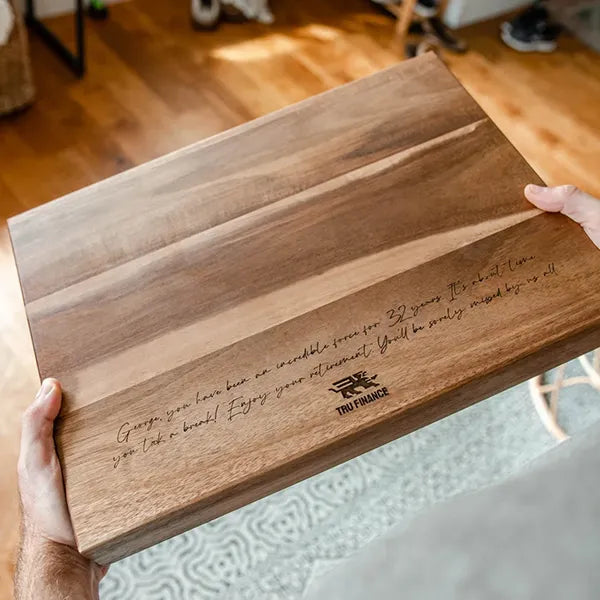A person holds a rectangular wooden cutting board with engraved text and a small 'TIN TRUCK' logo, photographed in a real indoor setting with a wood floor and furniture visible in the background.