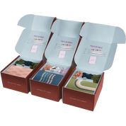 Three open gift boxes arranged side by side on a white background, each containing folded knit socks in different colors and patterns. The boxes are dark red-brown with pale blue inner flaps, and the socks include pastel pink, blue, green, and multicolor designs.