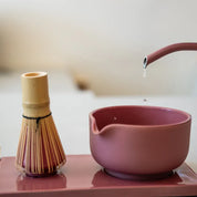 A pink Fellow ceramic tea bowl with a spout and a matching pink teapot pouring water, alongside a bamboo whisk on a pink tray, in a softly blurred indoor setting.