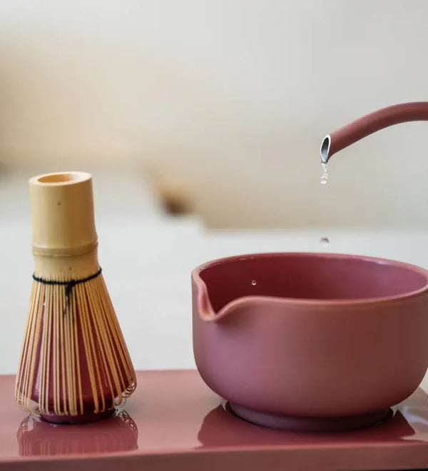 A pink Fellow ceramic tea bowl with a spout and a matching pink teapot pouring water, alongside a bamboo whisk on a pink tray, in a softly blurred indoor setting.