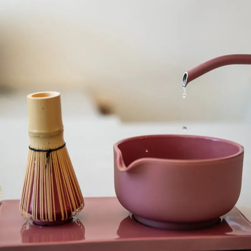 A pink Fellow ceramic tea bowl with a spout and a matching pink teapot pouring water, alongside a bamboo whisk on a pink tray, in a softly blurred indoor setting.