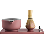 Fellow matcha tea set on a rectangular tray, featuring a dusty pink ceramic bowl with a metal lid, a bamboo whisk, and a small metal scoop on a solid white background.
