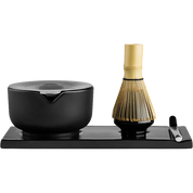 Fellow matcha tea set on a rectangular black tray, featuring a black ceramic bowl with a pouring spout, a bamboo whisk with a gold-toned handle, and a small black spoon on a white background.