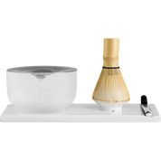 Fellow  matcha tea preparation set on a white rectangular base, featuring a frosted white ceramic bowl with a metallic silver lid, a bamboo whisk with a gold-colored handle, and a small black-handled tool on the right.