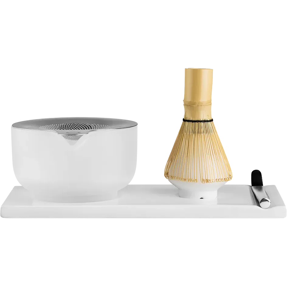 Fellow  matcha tea preparation set on a white rectangular base, featuring a frosted white ceramic bowl with a metallic silver lid, a bamboo whisk with a gold-colored handle, and a small black-handled tool on the right.