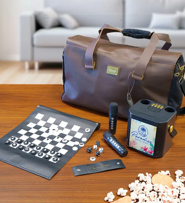 A brown leather-look duffel bag sits on a wooden table with a game set and party accessories arranged in front, including a checkers board, microphone, speaker, popcorn, and a small card. A blurred living room with a sofa is visible in the background.