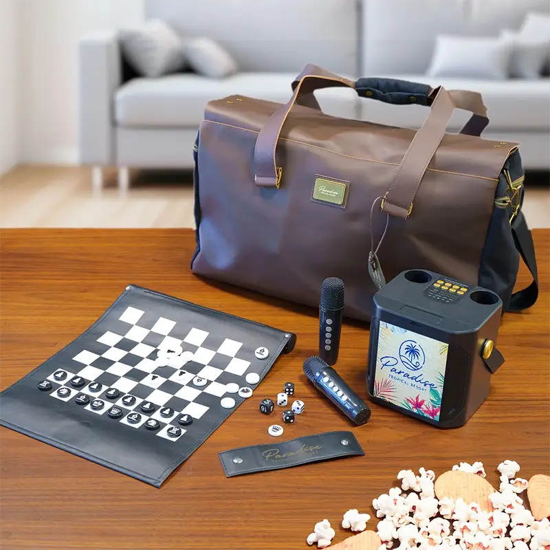 A brown leather-look duffel bag sits on a wooden table with a game set and party accessories arranged in front, including a checkers board, microphone, speaker, popcorn, and a small card. A blurred living room with a sofa is visible in the background.