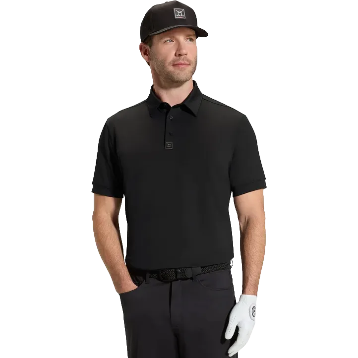 A man modeling a black short-sleeve polo shirt with a small square logo patch on the chest, paired with a black cap, dark pants, and a white golf glove, against a solid white background.