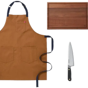 A brown canvas apron with dark navy straps and pockets, shown alongside a wooden cutting board and a chef's knife on a white background.