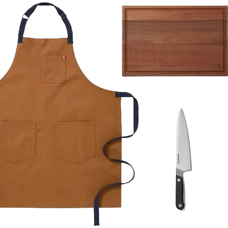 A brown canvas apron with dark navy straps and pockets, shown alongside a wooden cutting board and a chef's knife on a white background.