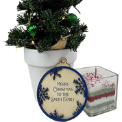 A round Christmas ornament with a blue border and the text 'Merry Christmas to the Smith Family' displayed in front of a small potted decorated tree, next to a square layered candle in a glass container.