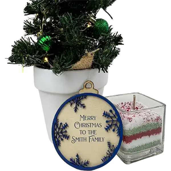 A round Christmas ornament with a blue border and the text 'Merry Christmas to the Smith Family' displayed in front of a small potted decorated tree, next to a square layered candle in a glass container.