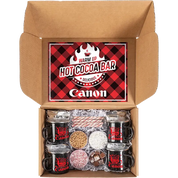 An open cardboard gift box containing a hot cocoa bar set. The lid features a red and black plaid graphic with the text 'WARM UP HOT COCOA BAR' and the brand name 'Canon'.
