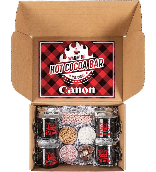 An open cardboard gift box containing a hot cocoa bar set. The lid features a red and black plaid graphic with the text 'WARM UP HOT COCOA BAR' and the brand name 'Canon'.
