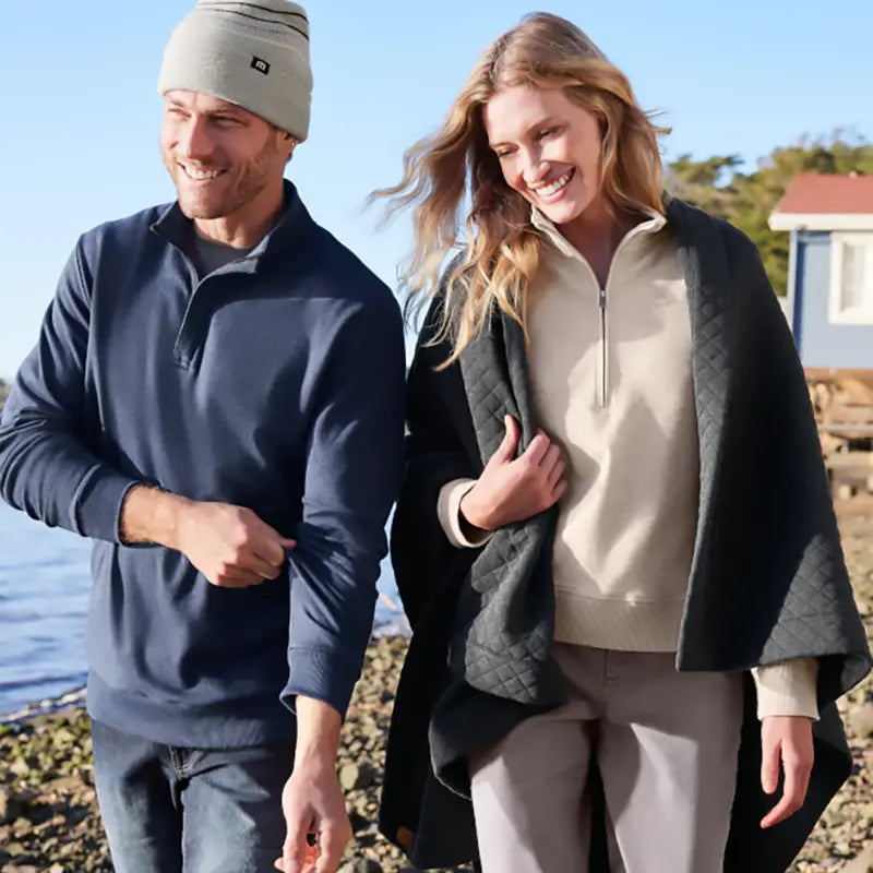 A man and a woman walking outdoors by the water wearing matching navy quarter-zip pullovers and light beige outerwear, with a blue sky and shoreline in the background.
