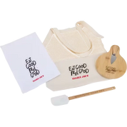 A Trader Joe's gift set featuring a cream mesh tote bag, a white dish towel, a wooden spatula, and a wooden cheese knife, all printed with the 'Eat Good Feel Good Trader Joe's' logo.