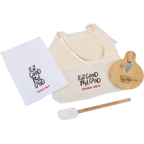 A Trader Joe's gift set featuring a cream mesh tote bag, a white dish towel, a wooden spatula, and a wooden cheese knife, all printed with the 'Eat Good Feel Good Trader Joe's' logo.