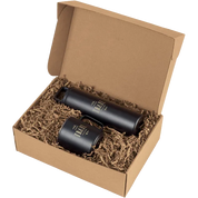An open kraft cardboard gift box filled with brown crinkle paper, containing two matte black insulated tumblers with gold foil branding that reads 'The Trail'.