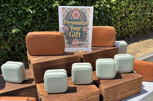A set of small zippered pouches and rectangular cases in tan and pale mint green, arranged on wooden display blocks outdoors with a green hedge in the background. A sign reading 'Personalize Your Gift' is visible behind the items.
