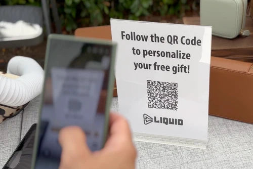 A hand holds a smartphone in front of a clear acrylic sign that reads 'Follow the QR Code to personalize your free gift!' with the LIQUID logo and a QR code, set on a table with a neutral background.