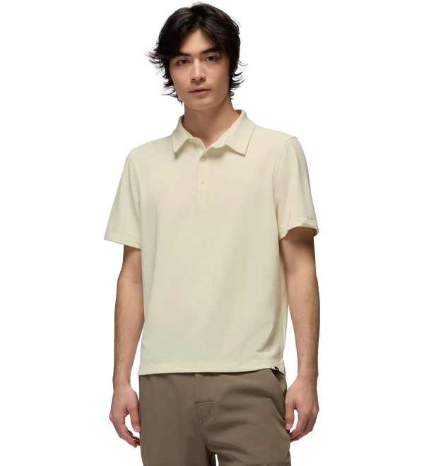A man wearing a light cream-colored short-sleeve polo shirt and brown pants, standing against a solid white background.