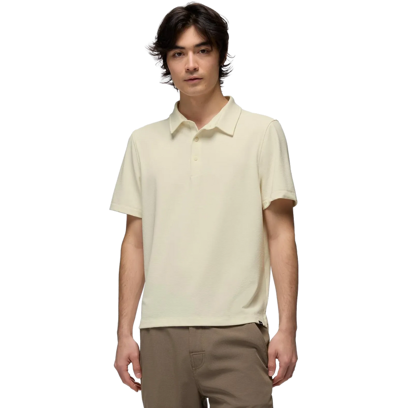 A man wearing a light cream-colored short-sleeve polo shirt and brown pants, standing against a solid white background.