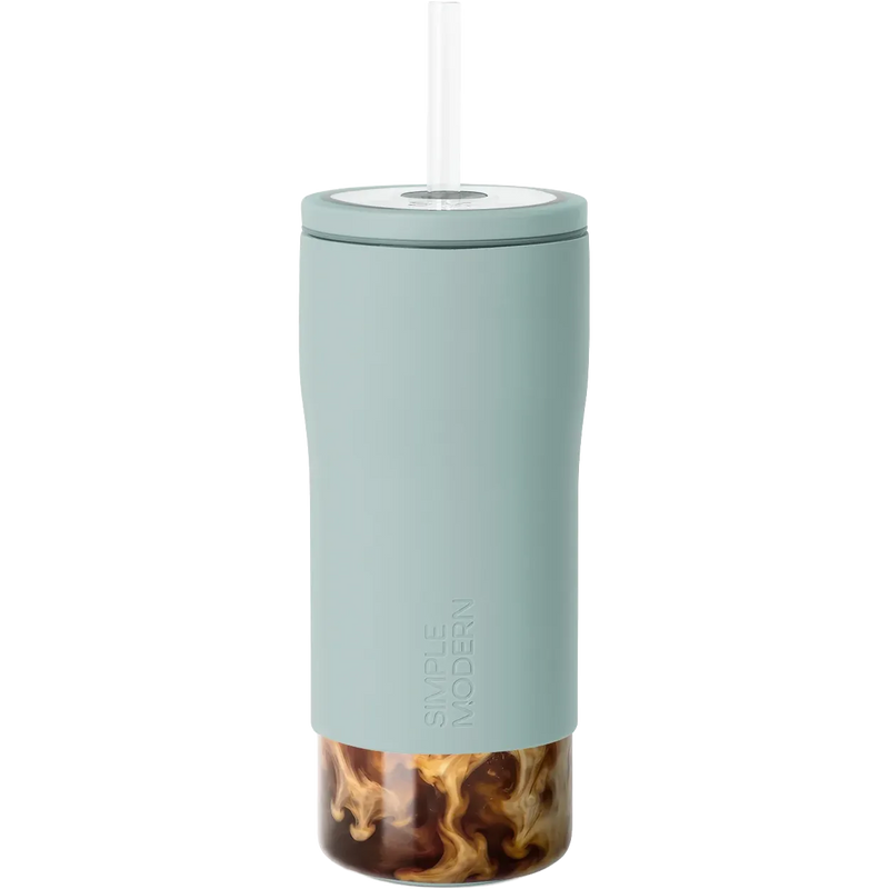 A tall, slim travel tumbler in a muted sage green color with a clear lid and straw, shown on a solid white background. The bottom portion of the tumbler is transparent, revealing a swirl of iced coffee inside.