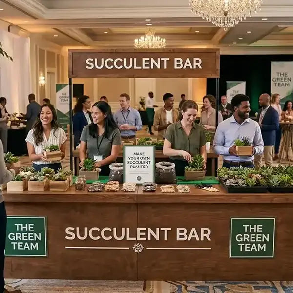 A bustling indoor event booth labeled 'SUCCULENT BAR' with people standing behind and around it, surrounded by potted succulents, small plants, and decorative items on a wooden counter. Signs for 'THE GREEN TEAM' are visible on the sides.