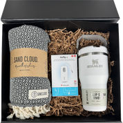 A gift set in a black box containing a patterned black and white beach towel from Sand Cloud, a white Stanley flip straw tumbler, and an AirFly earbud charging cable nestled in brown paper shred.