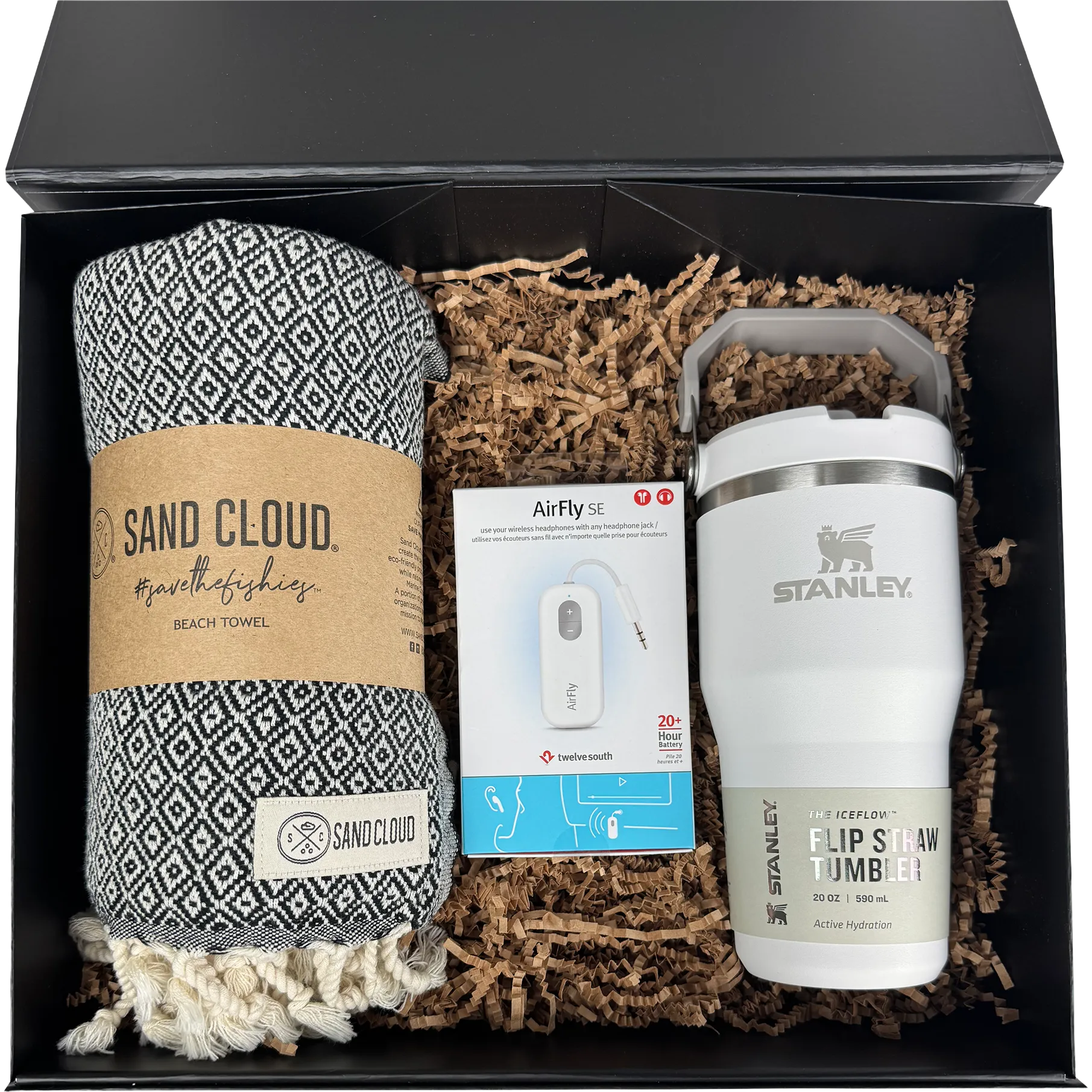 A gift set in a black box containing a patterned black and white beach towel from Sand Cloud, a white Stanley flip straw tumbler, and an AirFly earbud charging cable nestled in brown paper shred.