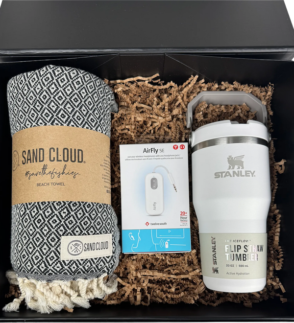 A gift set in a black box containing a patterned black and white beach towel from Sand Cloud, a white Stanley flip straw tumbler, and an AirFly earbud charging cable nestled in brown paper shred.
