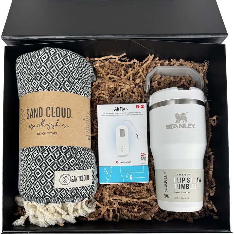 A gift set in a black box containing a patterned black and white beach towel from Sand Cloud, a white Stanley flip straw tumbler, and an AirFly earbud charging cable nestled in brown paper shred.